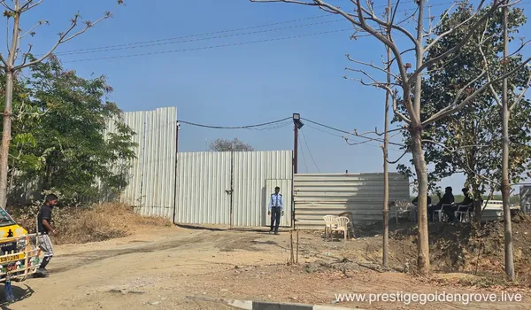 Site entrance with metal-sheet perimeter fencing and a security guard at the Prestige Golden Grove project, Velimela.